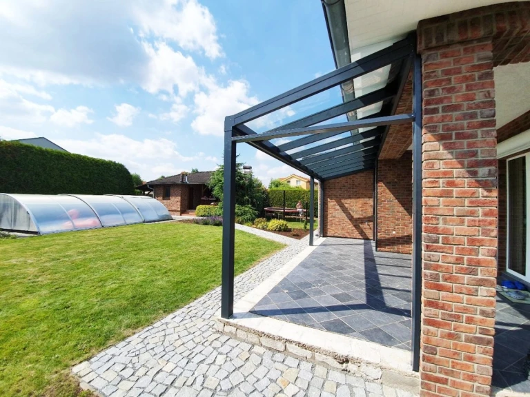 Eine moderne anthrazitfarbene Aluminium-Pergola mit Glasdach neben einem Backsteinhaus mit Garten und Swimmingpool.