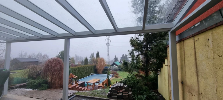 Blick von unterhalb der Aluminium-Pergola auf den Garten mit Swimmingpool an einem regnerischen Tag