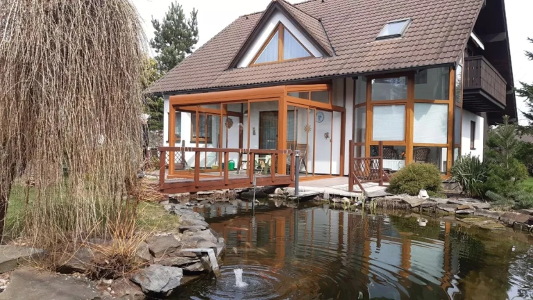 Eine verglaste Aluminium-Pergola neben einem Einfamilienhaus mit einem Gartenteich und einer Weide.
