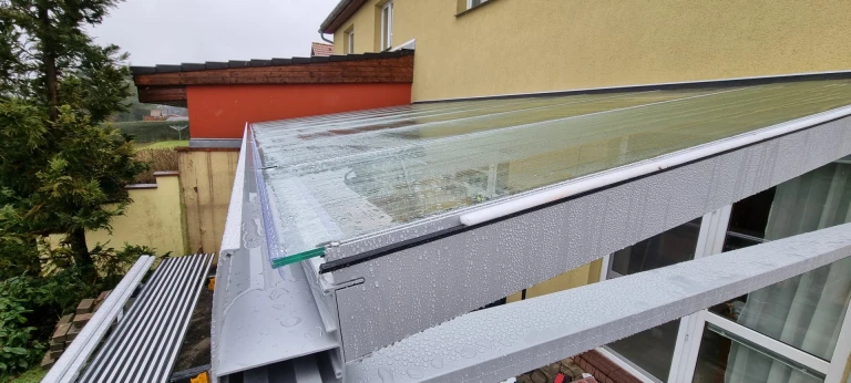 Ein Detailblick auf die Überdachung einer Aluminium-Pergola mit Glasfüllung, auf der Regentropfen liegen.