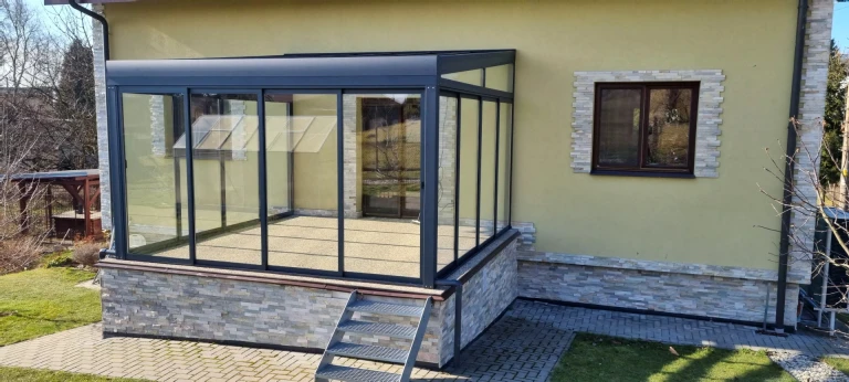 Verglaste Aluminium-Pergola vom Typ ROBUST, die an ein Einfamilienhaus angebaut ist und über eine kleine Treppe verfügt