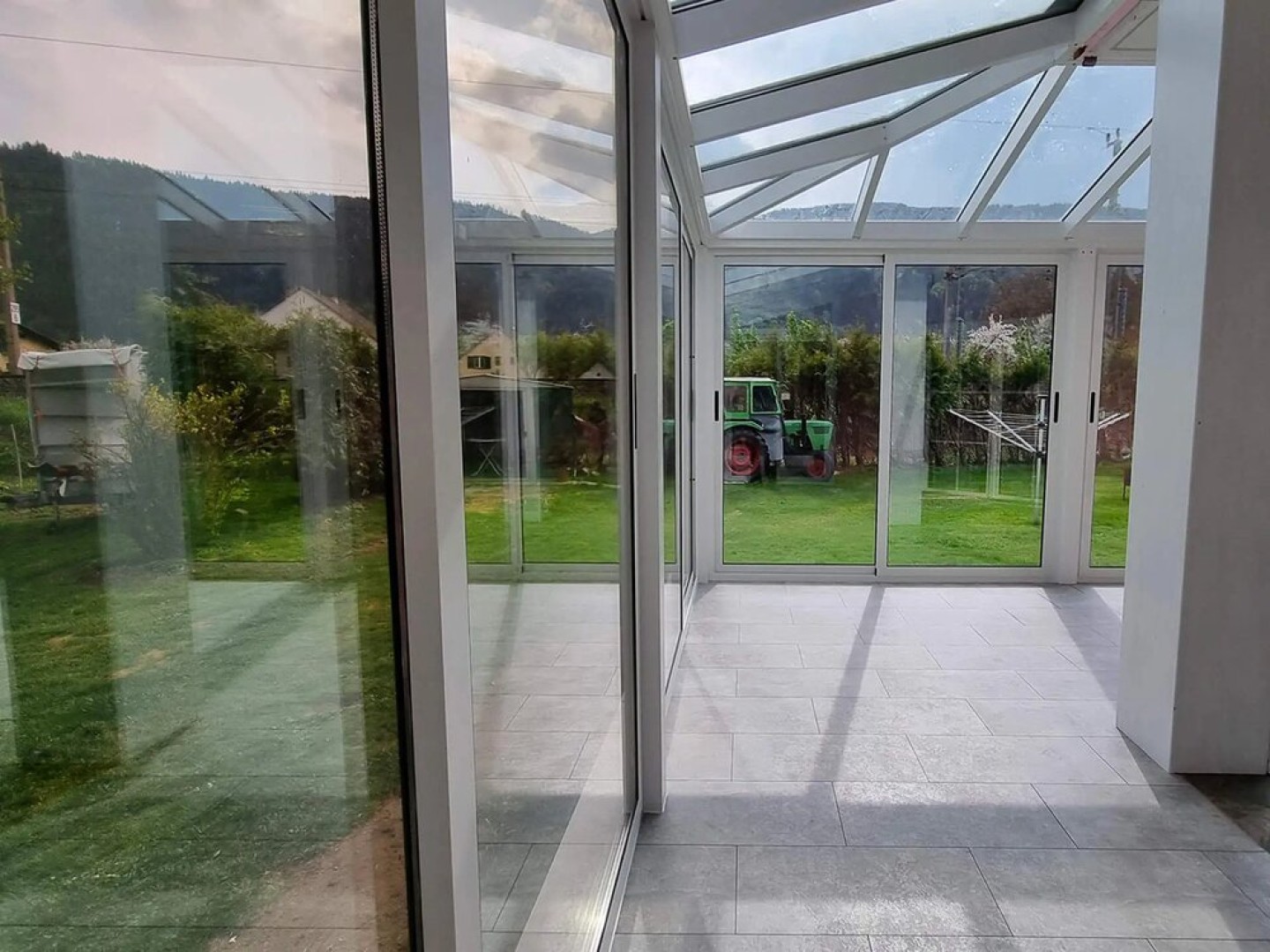 Blick vom Innenraum auf die verglaste Terrasse mit Garten und einem Traktor im Hintergrund