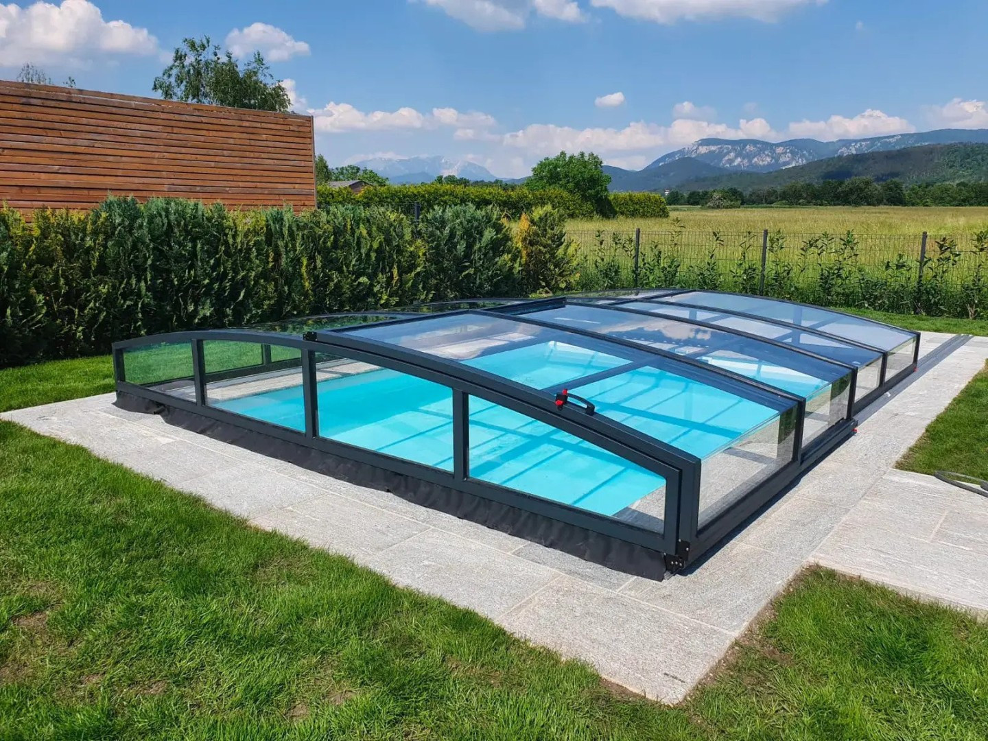 Eine moderne, niedrige STAR-Schwimmbadüberdachung in Anthrazit, die in einem Garten mit Blick auf die Berge steht