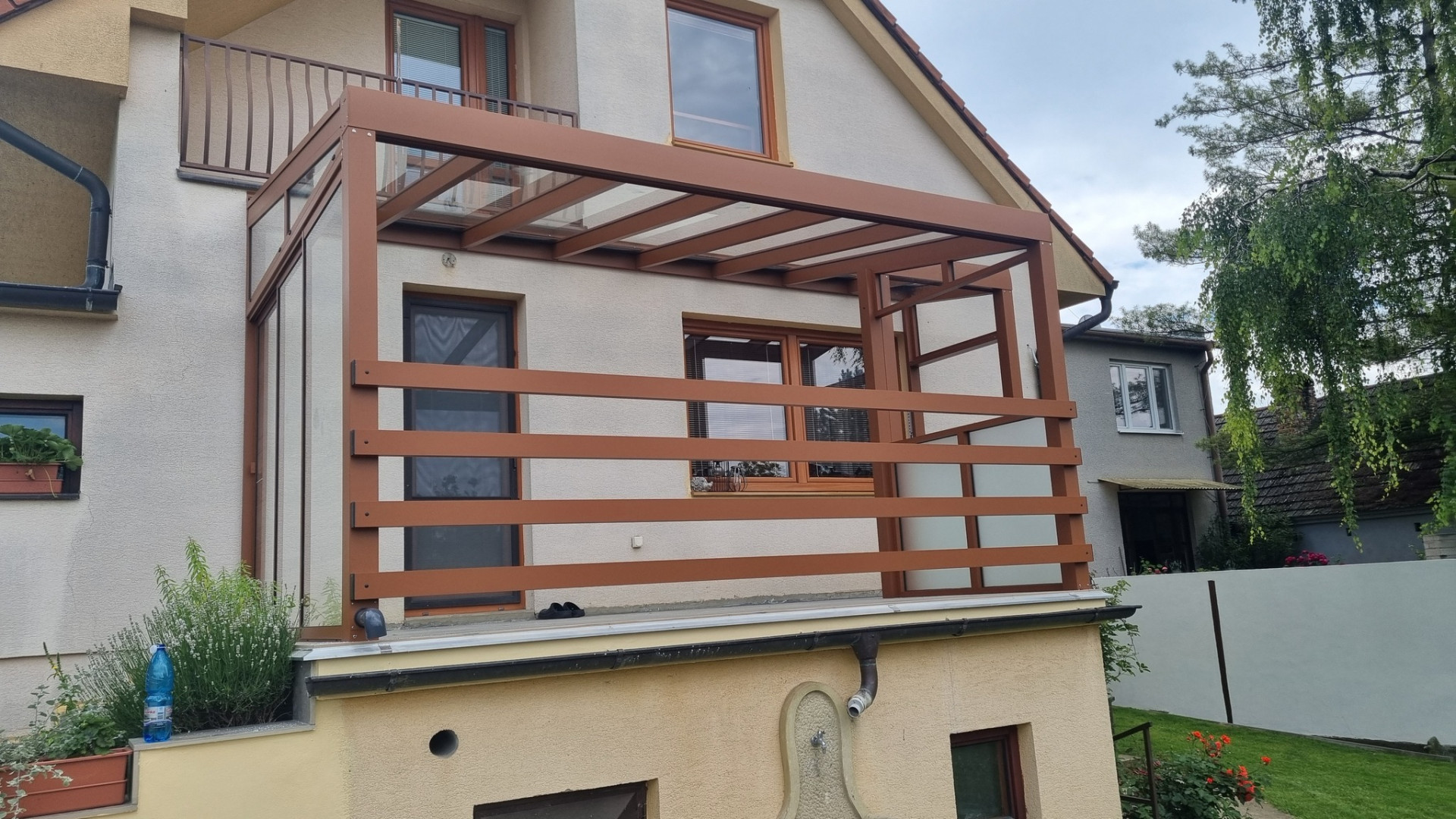 Eine braune, ungewöhnlich gestaltete Aluminium-Pergola mit Glasdach und seitlichem Sonnenschutz auf dem Balkon eines Einfamilienhauses