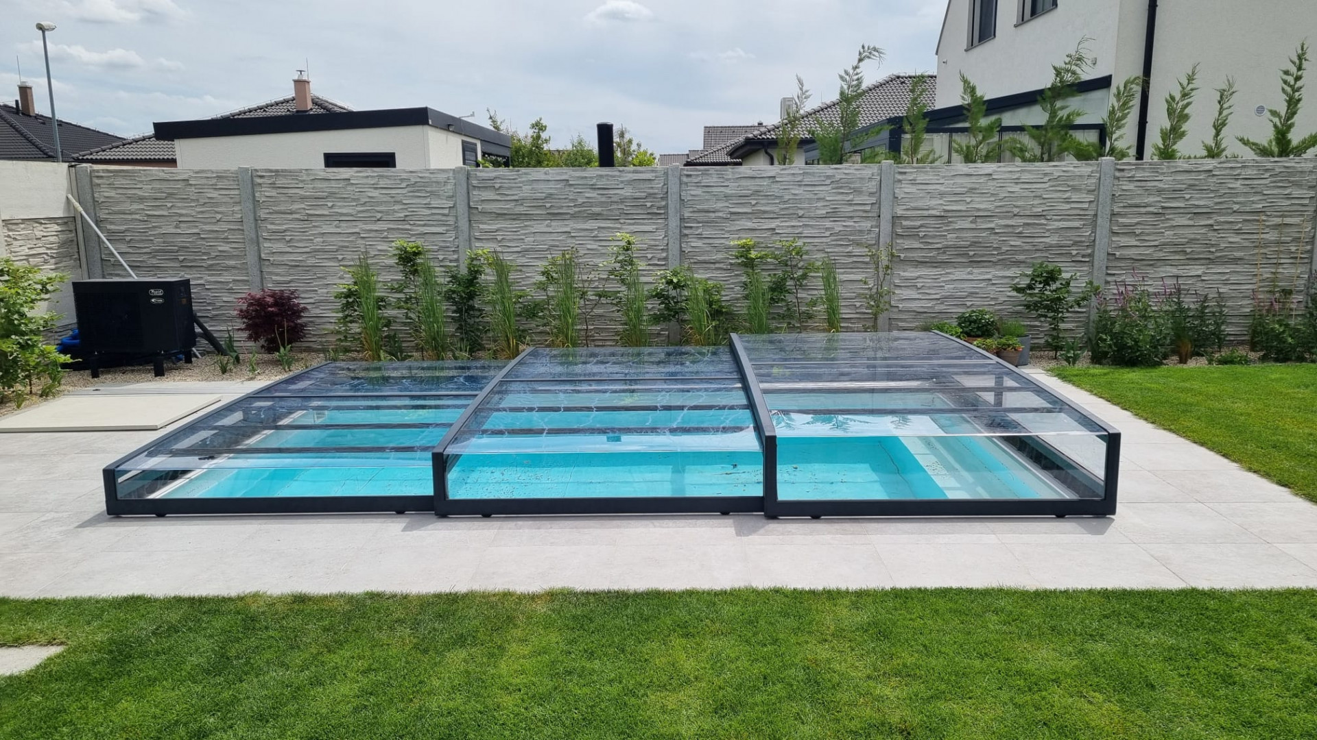 Eine moderne, niedrige Schwimmbadüberdachung mit anthrazitfarbener Konstruktion und transparentem Füllmaterial im Garten eines Einfamilienhauses.