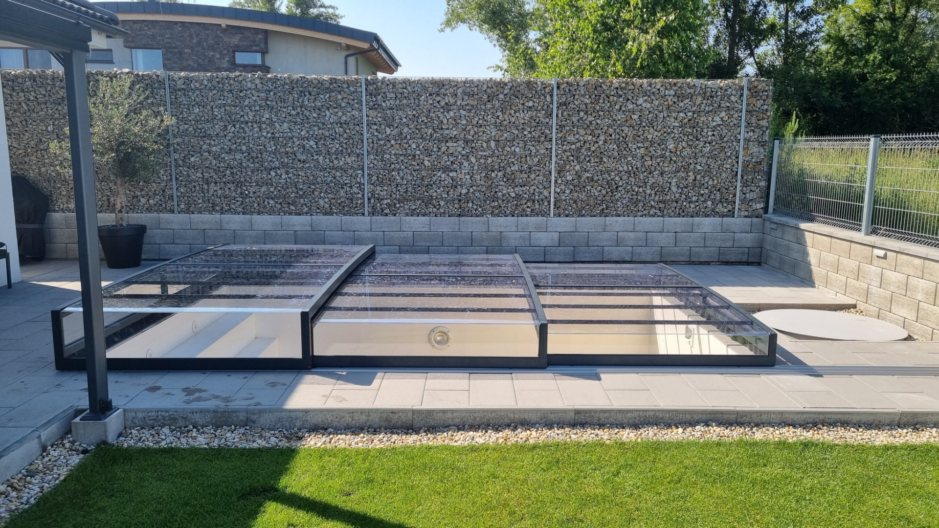 Niedrige Schwimmbadüberdachung EXCLUSIVE aus transparentem Polycarbonat im Garten neben einer Steinmauer.