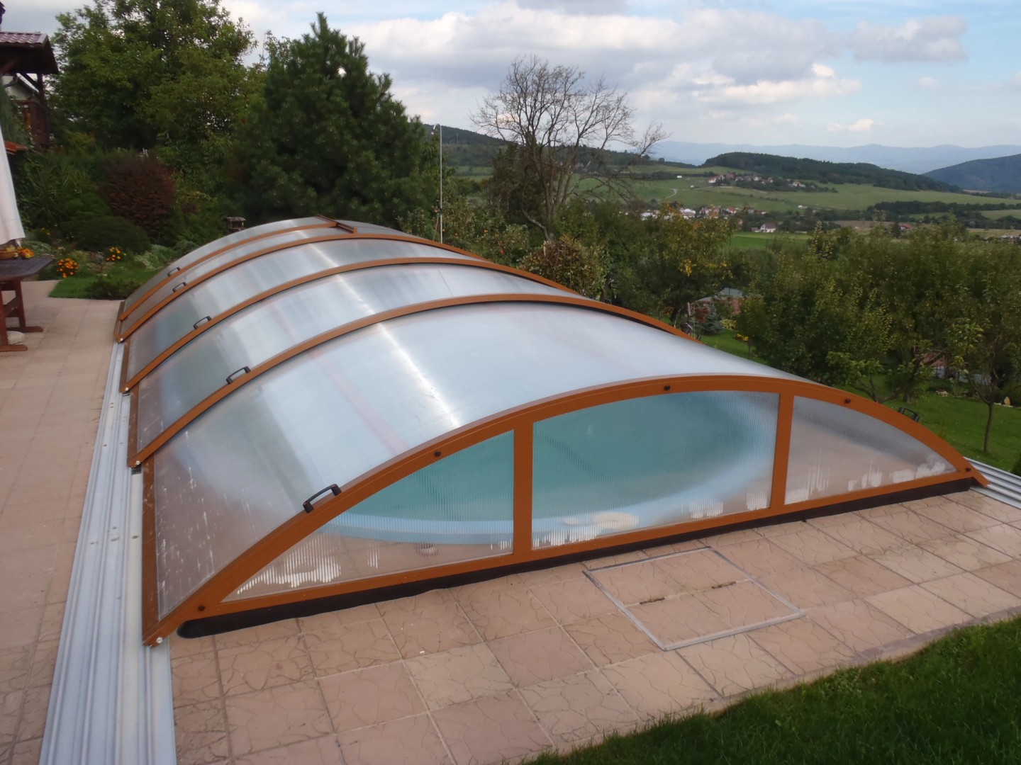 Eine geschlossene, niedrige, gewölbte Überdachung des Schwimmbads mit braunem Rahmen, die auf einer Terrasse mit Blick auf die hügelige Landschaft steht.