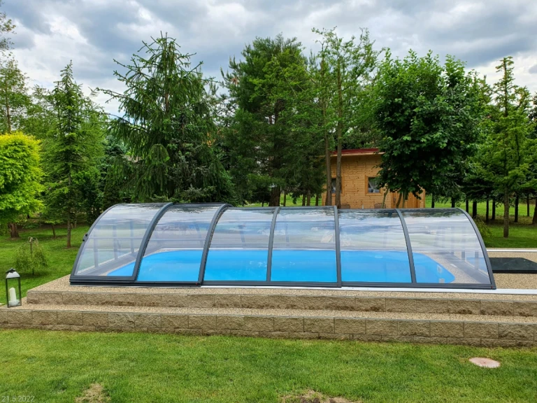 Eine geschlossene, bogenförmige, niedrige Schwimmbadüberdachung über einem blauen Pool in einem Garten mit Bäumen.