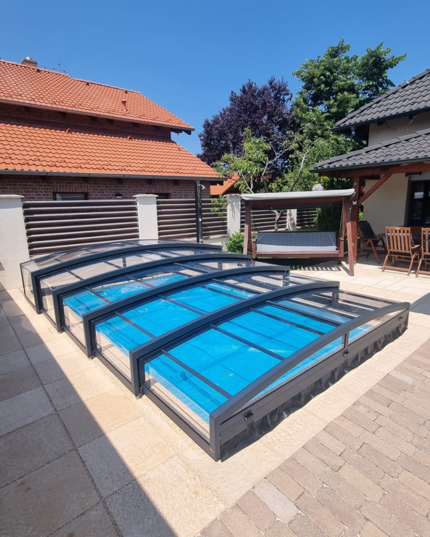 Eine niedrige, anthrazitfarbene Teleskop-Schwimmbadüberdachung auf dem gepflasterten Boden im Garten eines Einfamilienhauses.