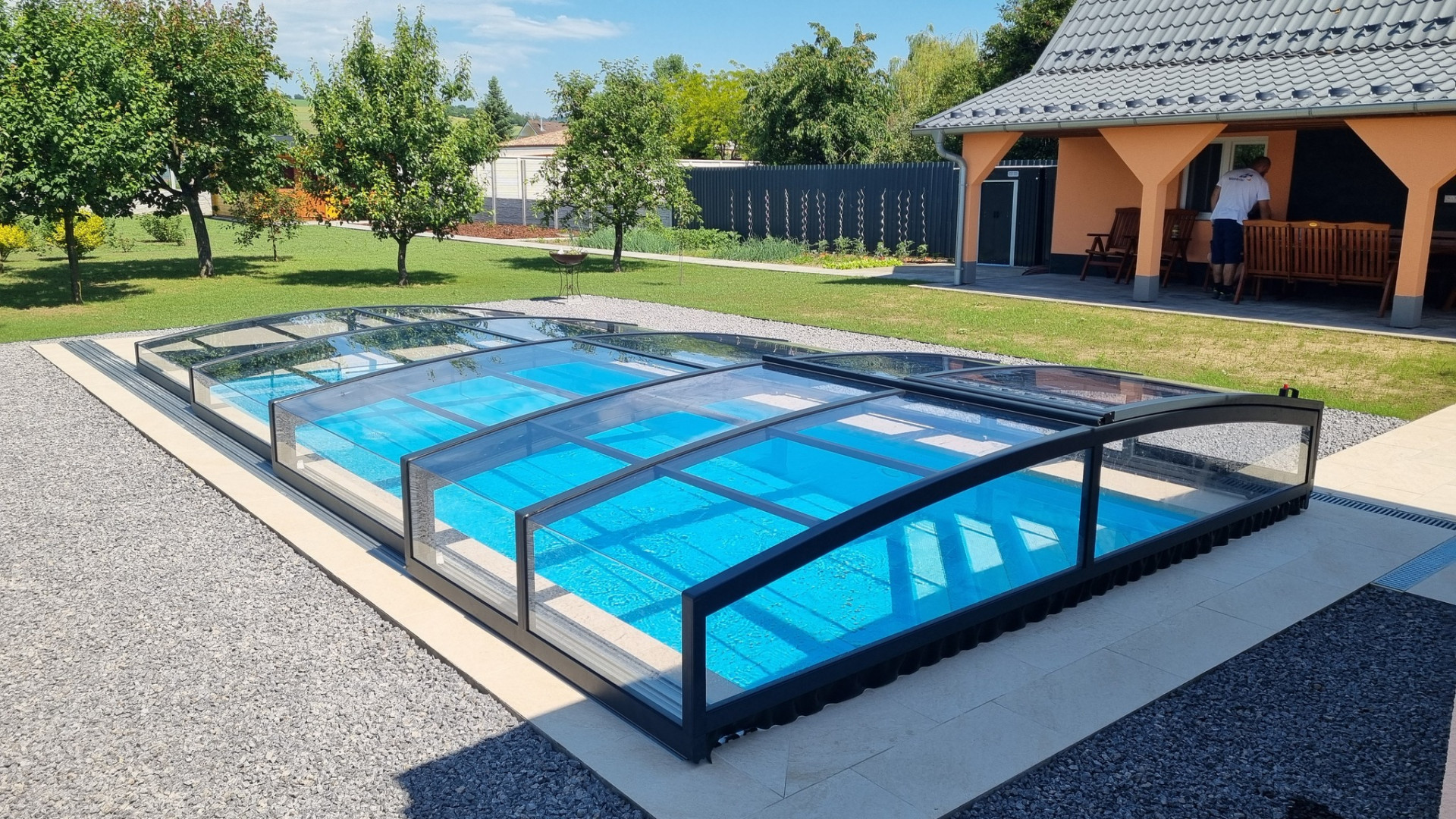 Eine moderne, niedrige PREMIER-Schwimmbadüberdachung mit dunklem Rahmen im Garten eines Einfamilienhauses.