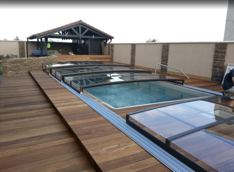 Eine moderne, niedrige Schwimmbadüberdachung mit verschiebbaren Glassegmenten auf der Holzterrasse eines Einfamilienhauses.