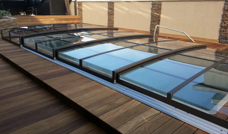 Eine niedrige, anthrazitfarbene Schwimmbadüberdachung mit transparenten Füllungen, die auf einer Holzterrasse errichtet wurde