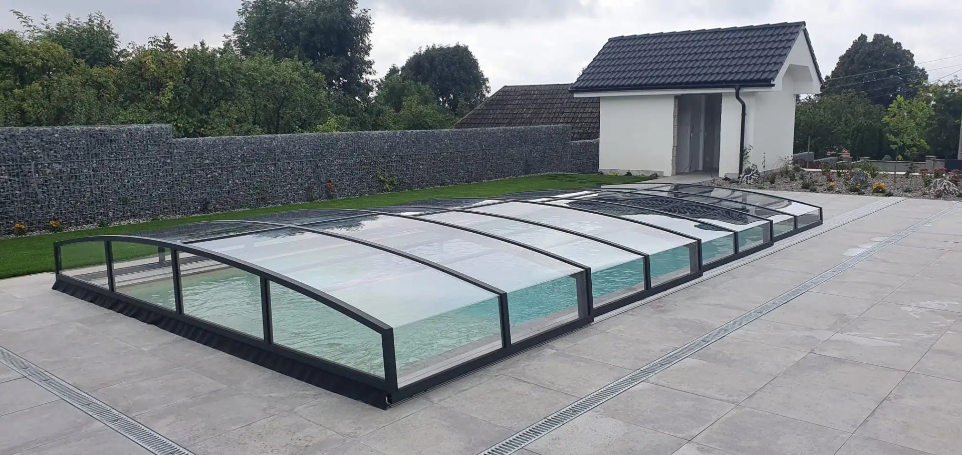 Eine moderne, niedrige Schwimmbadüberdachung mit schwarzer Konstruktion und klarem Glas auf der Terrasse neben dem Haus.