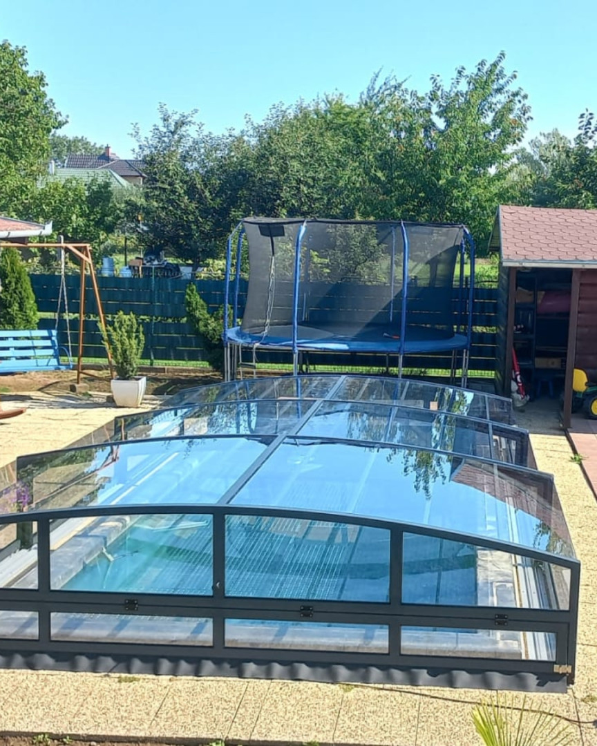 Moderne, niedrige Schwimmbadüberdachung mit anthrazitfarbenem Rahmen im Garten mit Trampolin