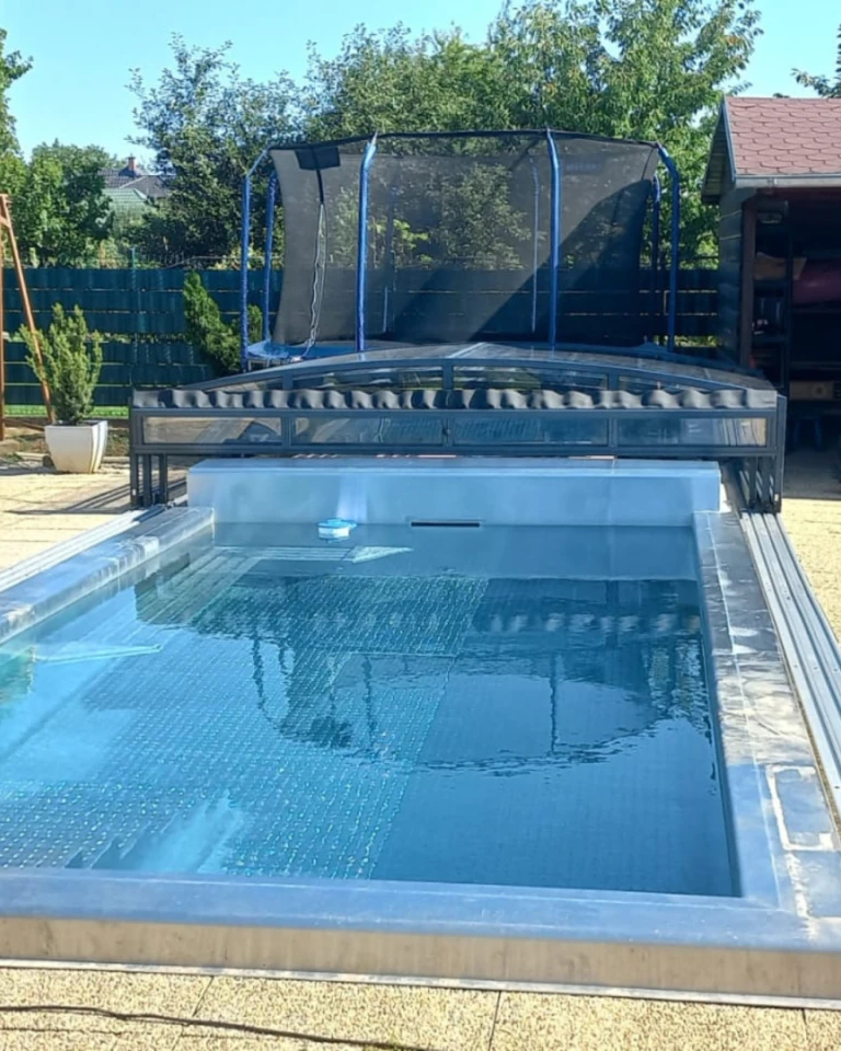 Ein modernes Schwimmbecken mit einer zurückversetzten, niedrigen Überdachung im Garten mit einem Trampolin.