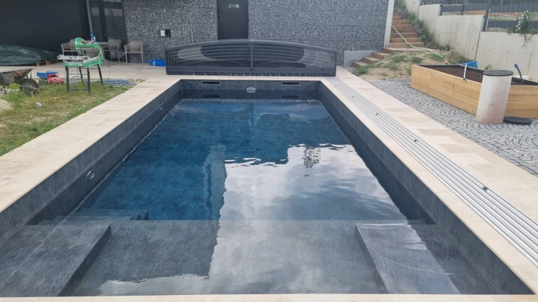 Ein moderner, in den Boden eingelassener Pool mit dunkler Verkleidung und einer zurückversetzten, niedrigen Überdachung auf der Terrasse neben dem Haus.