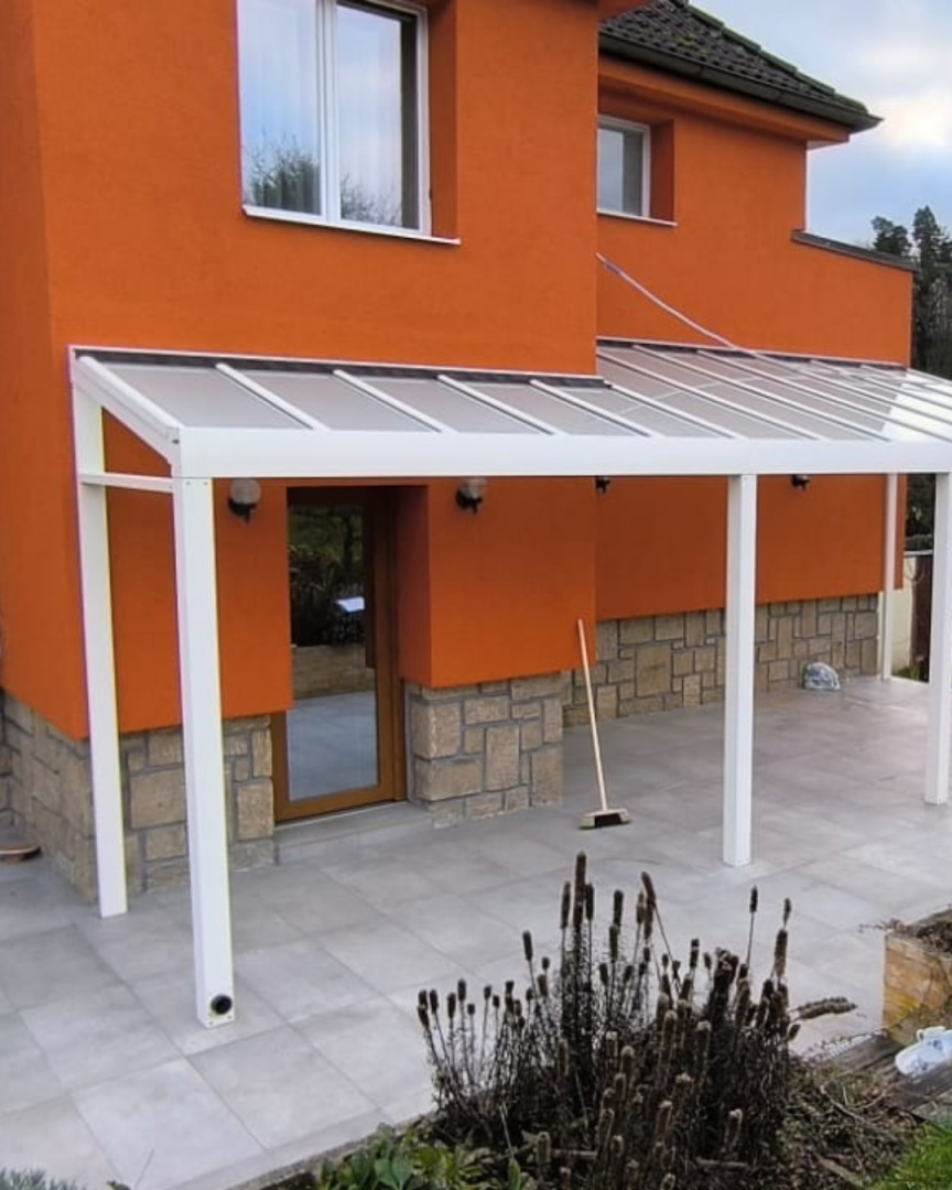Eine moderne weiße Aluminium-Pergola, die an einem orangefarbenen Haus mit Glasdach und Pflasterboden befestigt ist