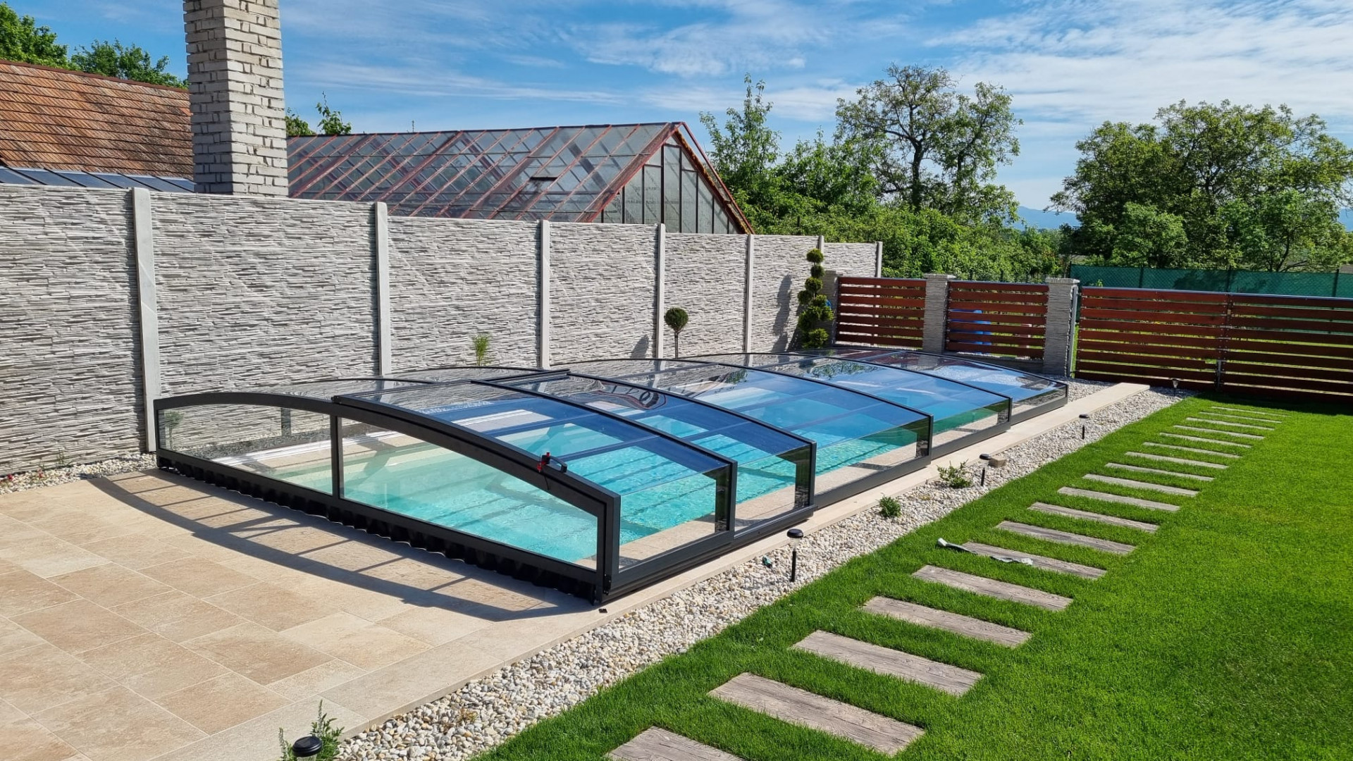 Eine moderne, niedrige Schwimmbadüberdachung mit anthrazitfarbener Konstruktion und klarem Glas in einem gepflegten Garten.