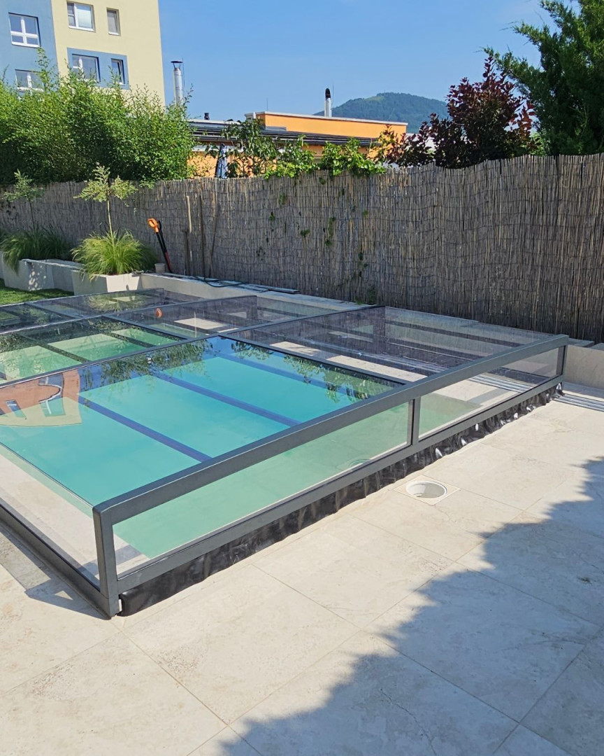 Eine moderne, niedrige Glasüberdachung für das Schwimmbecken mit anthrazitfarbenem Rahmen auf einer gepflasterten Terrasse.