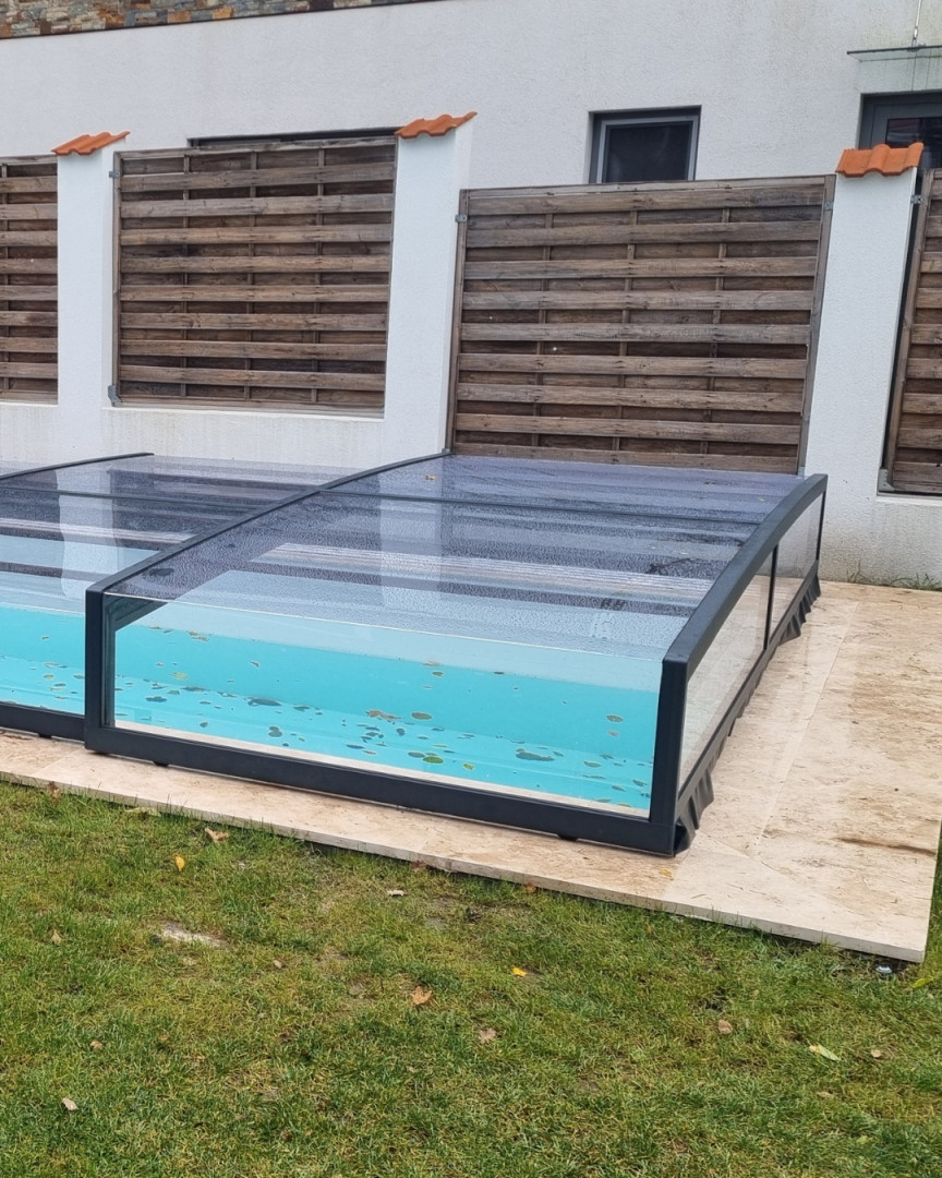 Eine moderne, niedrige Schwimmbadüberdachung mit anthrazitfarbenem Rahmen und transparentem Polycarbonat im Garten eines Einfamilienhauses.