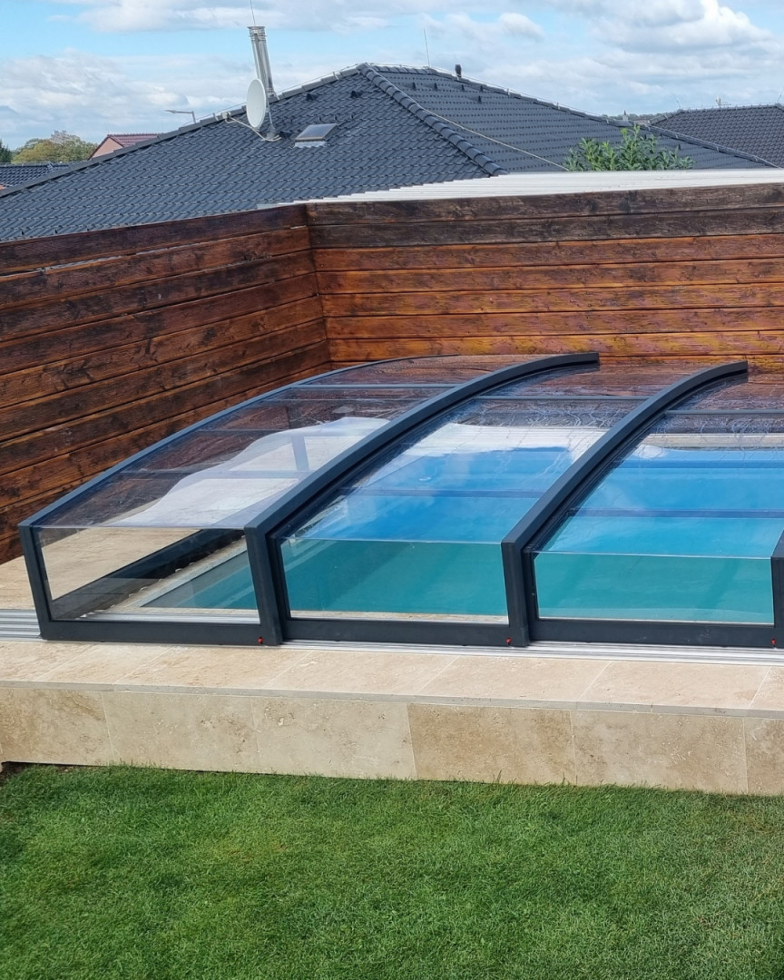 Moderne, niedrige Schwimmbadüberdachung mit anthrazitfarbenem Rahmen und transparentem Füllmaterial neben einem Holzzaun