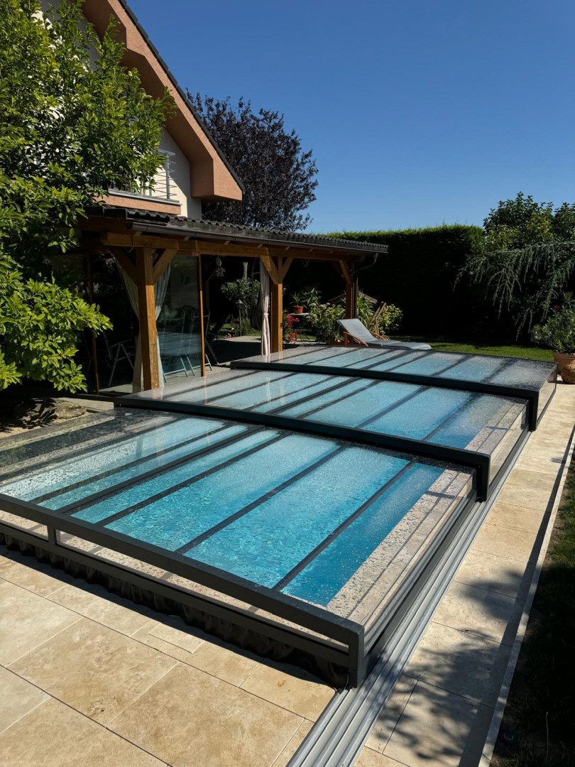 Moderne, niedrige Schwimmbadüberdachung mit anthrazitfarbener Konstruktion und transparenter Verkleidung im Garten eines Einfamilienhauses.