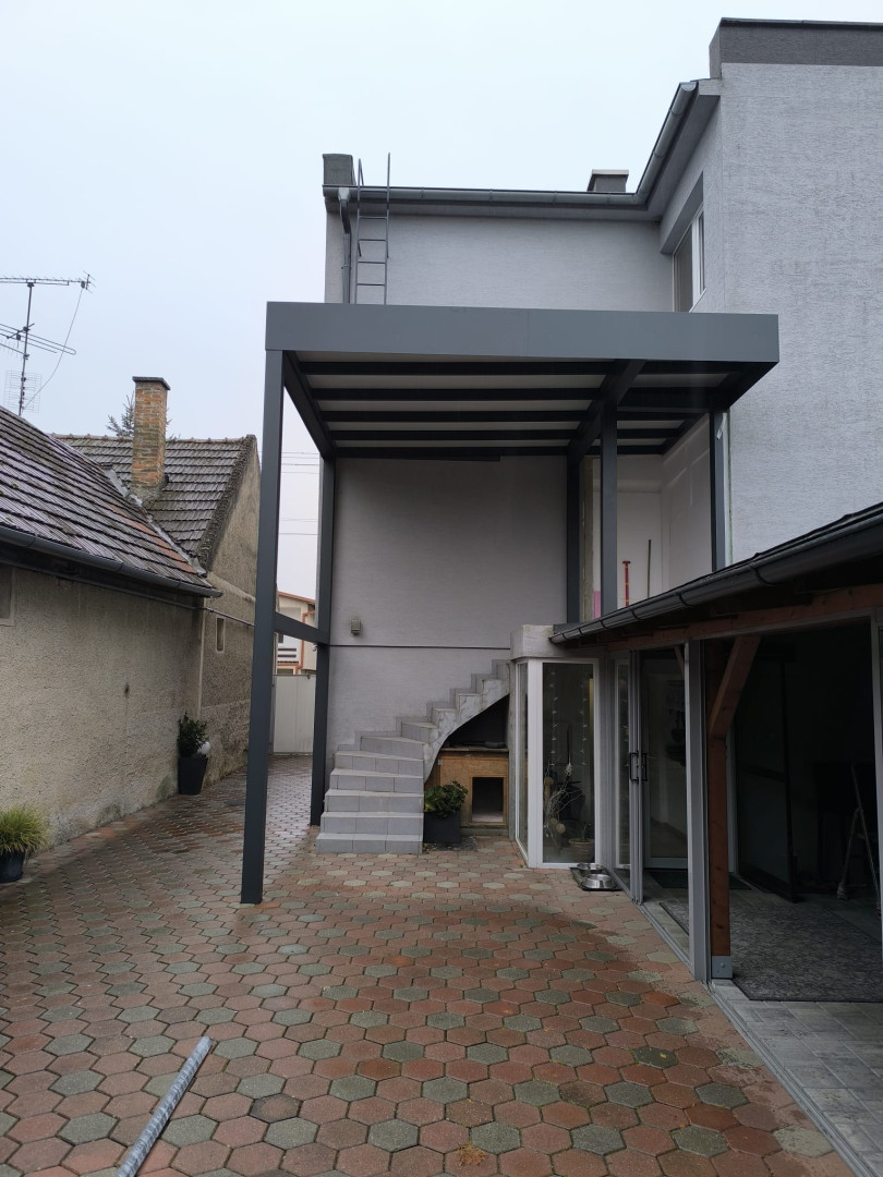 Eine moderne, maßgefertigte graue Aluminium-Pergola, die auf der Terrasse eines Einfamilienhauses über der Außentreppe installiert wurde.