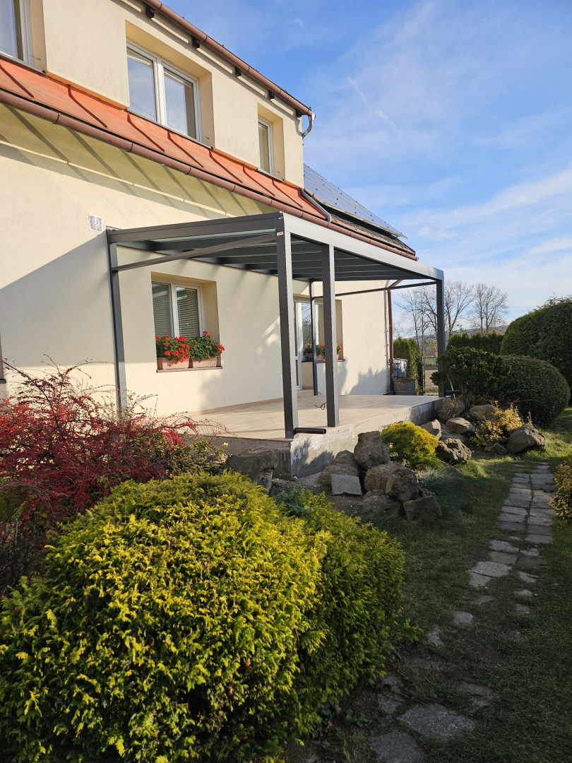 Eine moderne Aluminium-Pergola mit verglastem Dach, die an ein Einfamilienhaus mit Garten angebaut wurde.