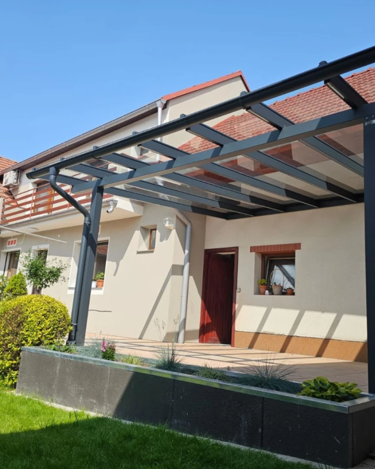 Eine moderne, anthrazitfarbene Aluminium-Pergola mit Glasdach, die an ein Einfamilienhaus angebaut ist