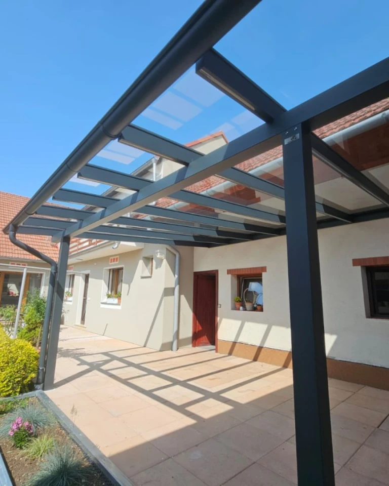 Eine moderne anthrazitfarbene Aluminium-Pergola mit Glasdach, die an ein Einfamilienhaus mit gepflasterter Terrasse angebaut ist.