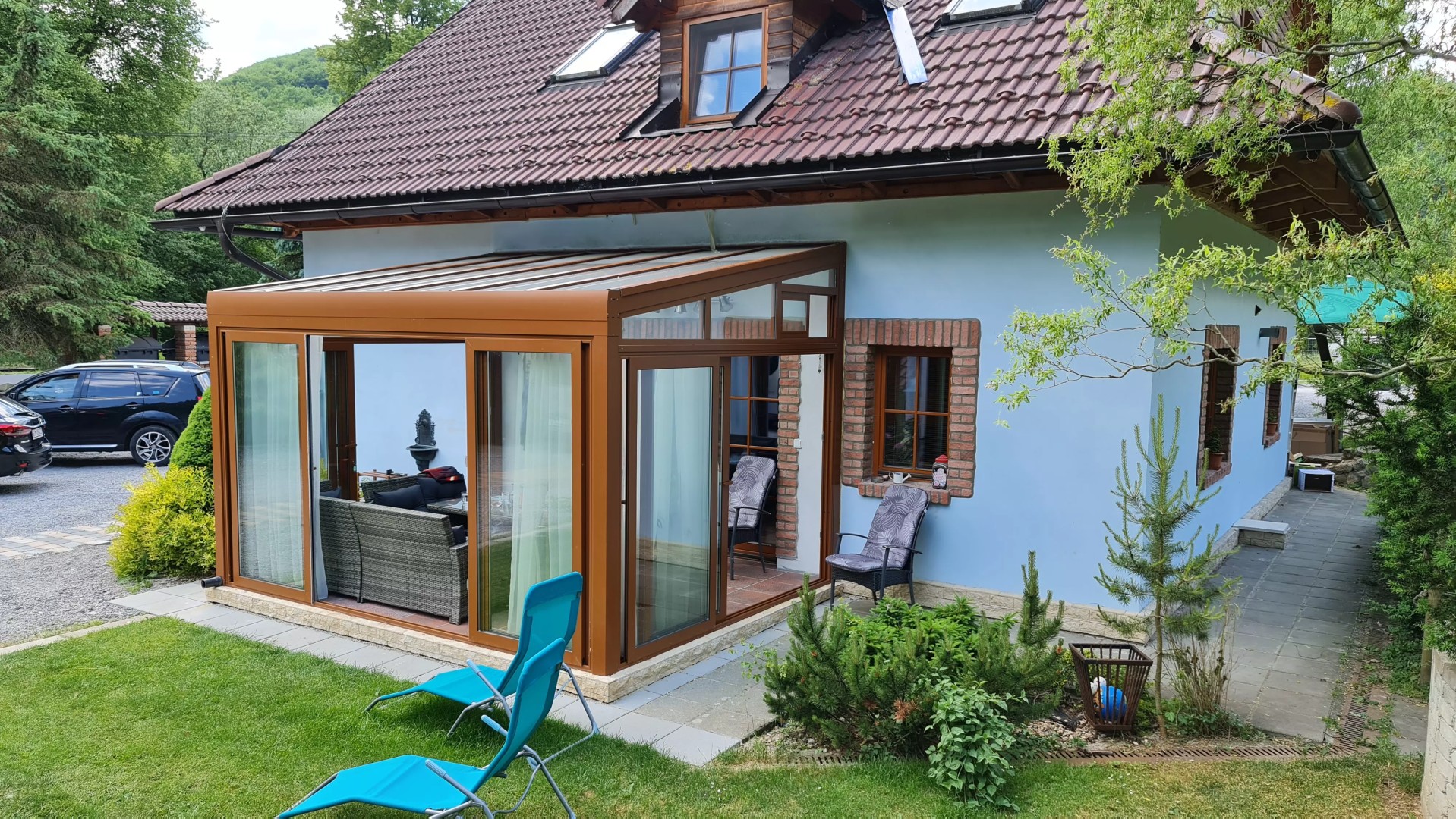Eine verglaste Aluminium-Pergola vom Typ ROBUST in brauner Farbe, die an ein Einfamilienhaus angebaut ist und mit Gartenmöbeln ausgestattet ist.