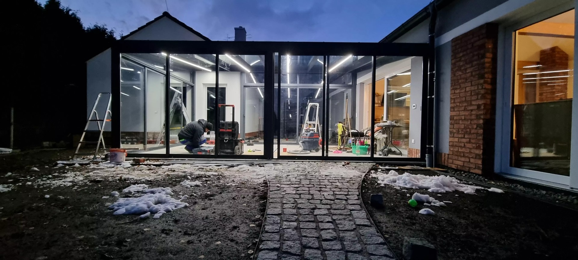 Abendlicher Blick auf die Montage einer verglasten Aluminium-Pergola mit integrierter LED-Beleuchtung an einem Einfamilienhaus.