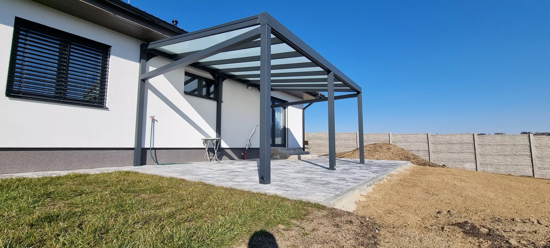 Eine moderne anthrazitfarbene Aluminium-Pergola mit Glasdach, die an ein Einfamilienhaus auf einer gepflasterten Terrasse angebaut ist.
