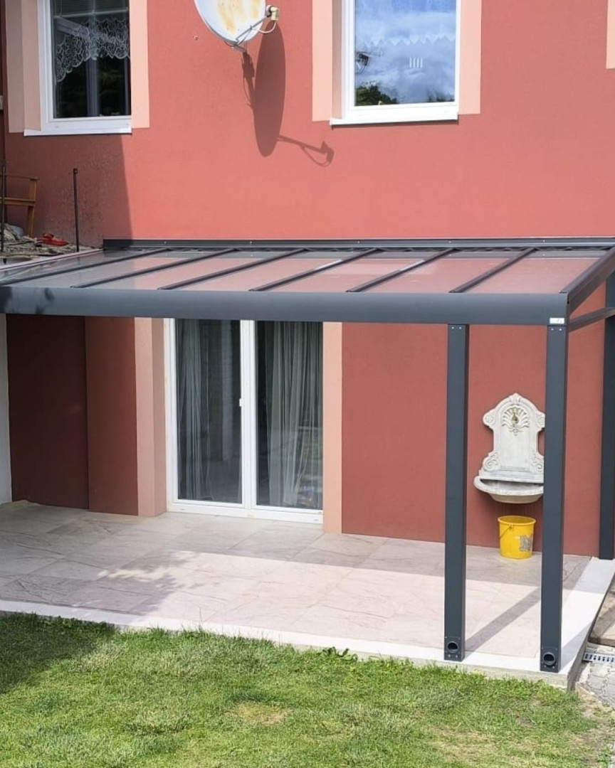 Eine moderne Aluminium-Pergola mit verglastem Dach, die an der Wand des roten Hauses über der Terrasse angebracht ist.