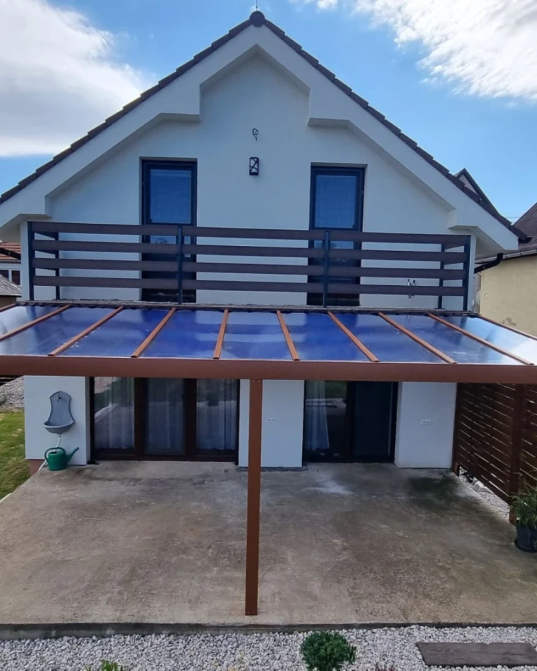Eine braune Aluminium-Pergola mit Glasdach, die an ein Einfamilienhaus mit Balkon angebaut ist.