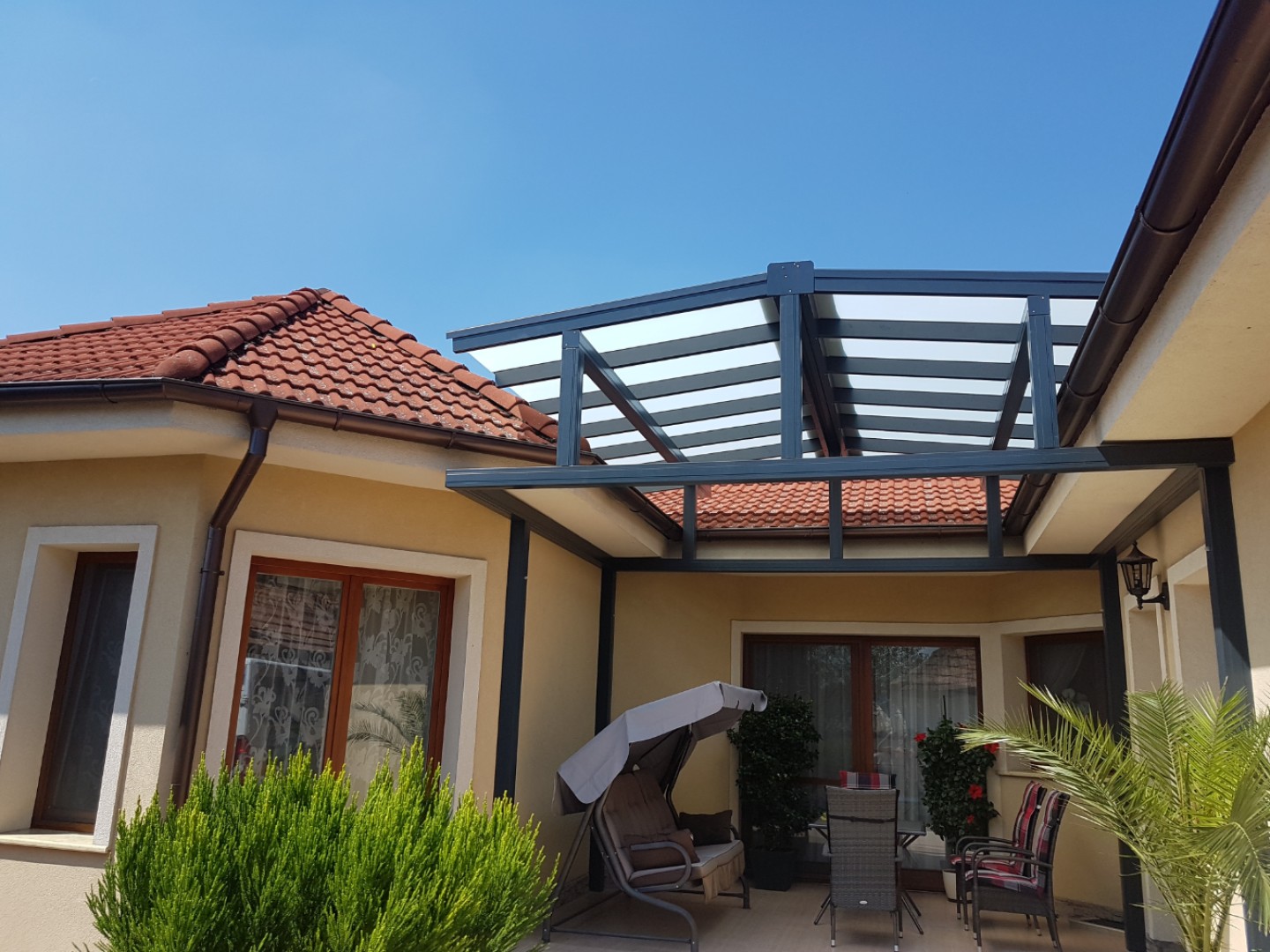 Moderne Aluminium-Pergola „ROBUST“ mit verglastem Dach, die an ein Einfamilienhaus angebaut ist, mit Terrassenmöbeln und einer Schaukel.