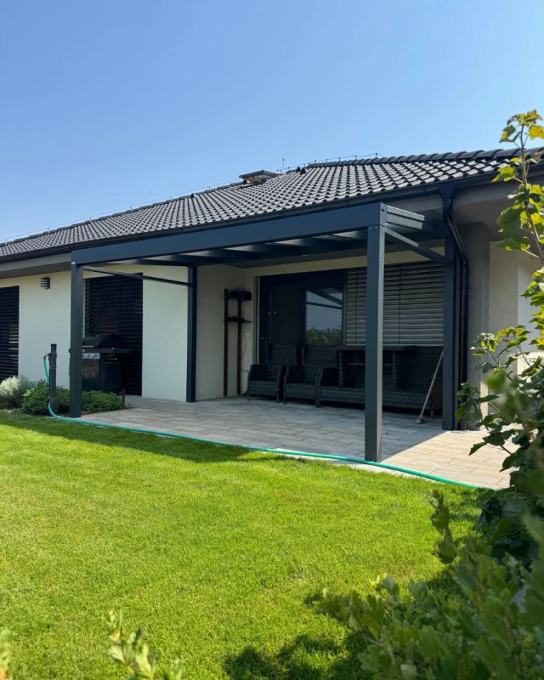 Eine moderne, dunkelgraue Aluminium-Pergola vom Typ „Robust“ auf der Terrasse eines Einfamilienhauses mit Gartenmöbeln und Rasenfläche