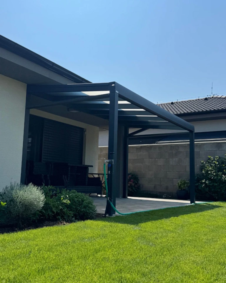 Eine moderne, dunkelgraue Aluminium-Pergola mit Glasdach, die an ein Einfamilienhaus mit Garten angebaut ist
