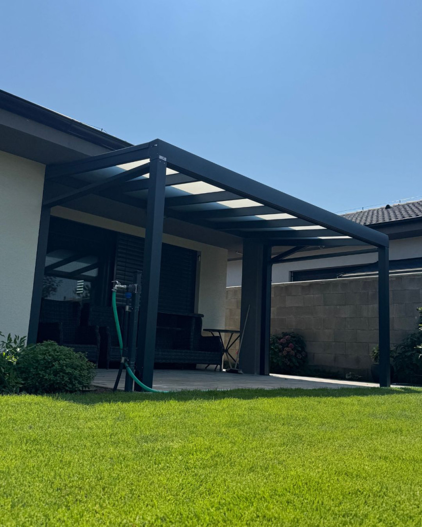 Eine moderne, dunkelgraue Aluminium-Pergola, die an das Haus angebaut ist, mit einem Glasdach und einem gepflegten Rasen