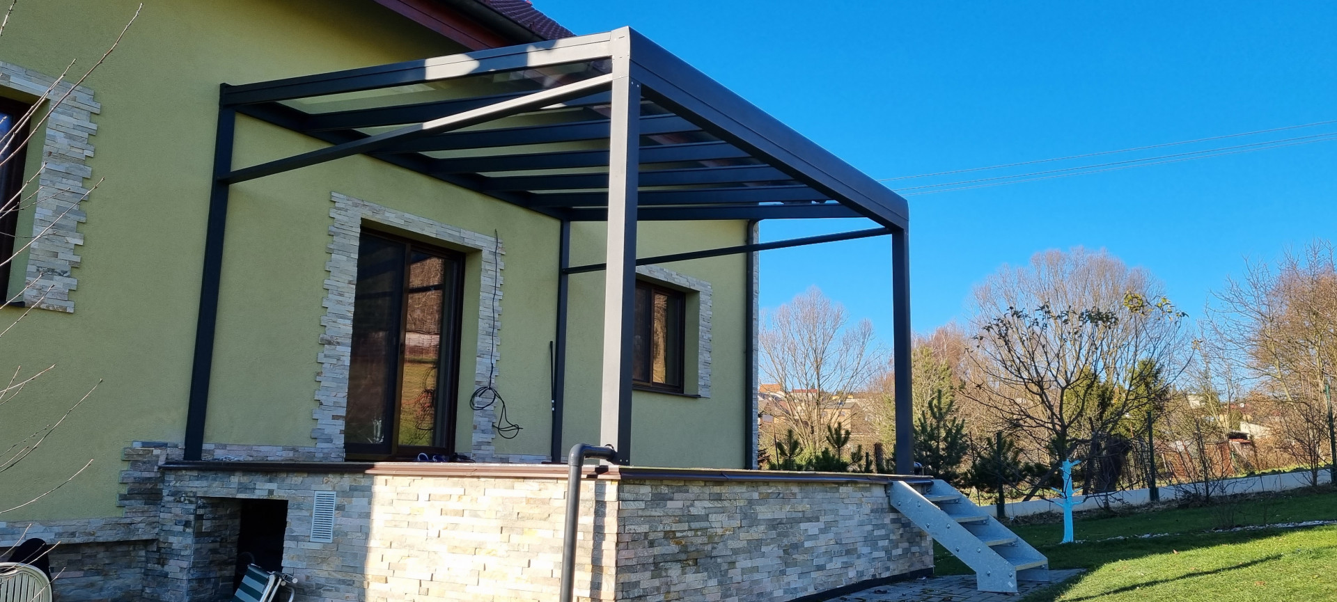 Eine moderne, dunkelgraue Aluminium-Pergola vom Typ ROBUST mit Glasdach, die auf der Terrasse eines Einfamilienhauses montiert wurde.