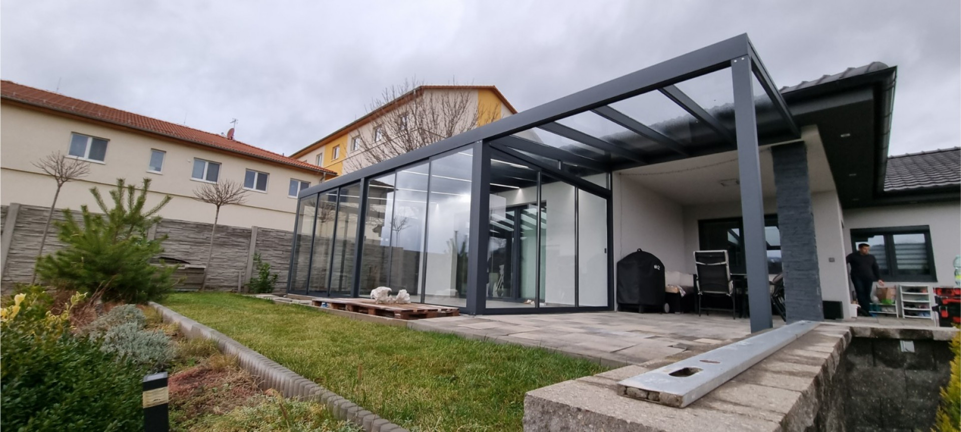 Eine moderne, verglaste Aluminium-Pergola mit anthrazitfarbener Konstruktion, die neben einem Einfamilienhaus in Klášterec errichtet wurde.