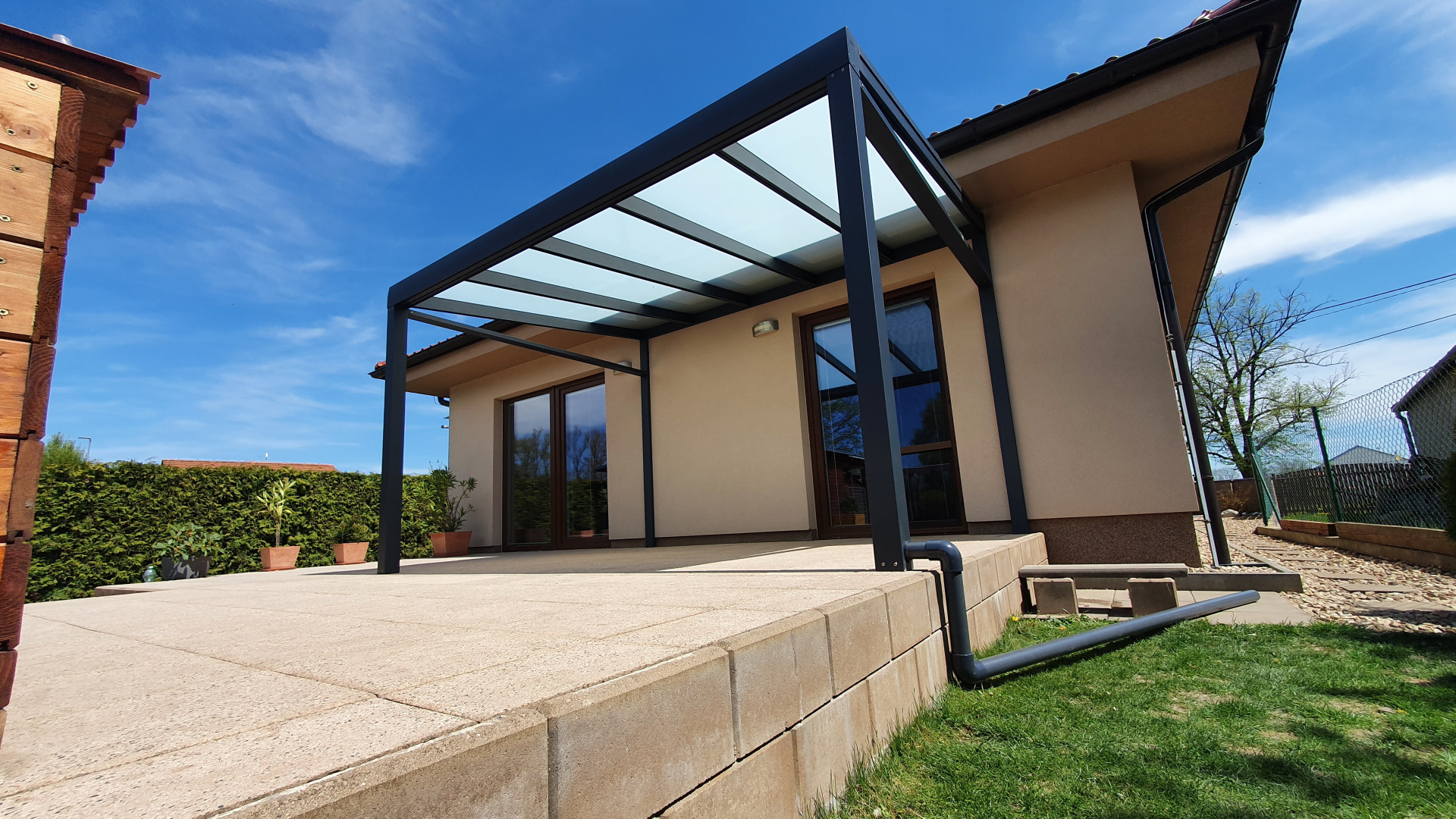 Eine moderne, dunkelgraue Aluminium-Pergola mit Glasdach, die an einem Einfamilienhaus auf einer gepflasterten Terrasse angebracht ist.