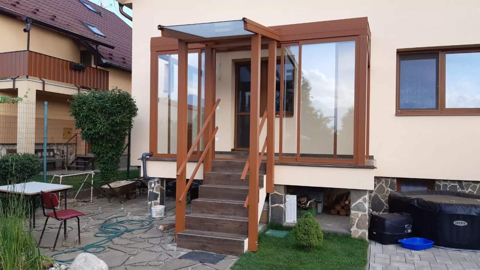 Eine braune, verglaste Aluminium-Pergola mit einem Vordach über der Eingangstreppe eines Einfamilienhauses.