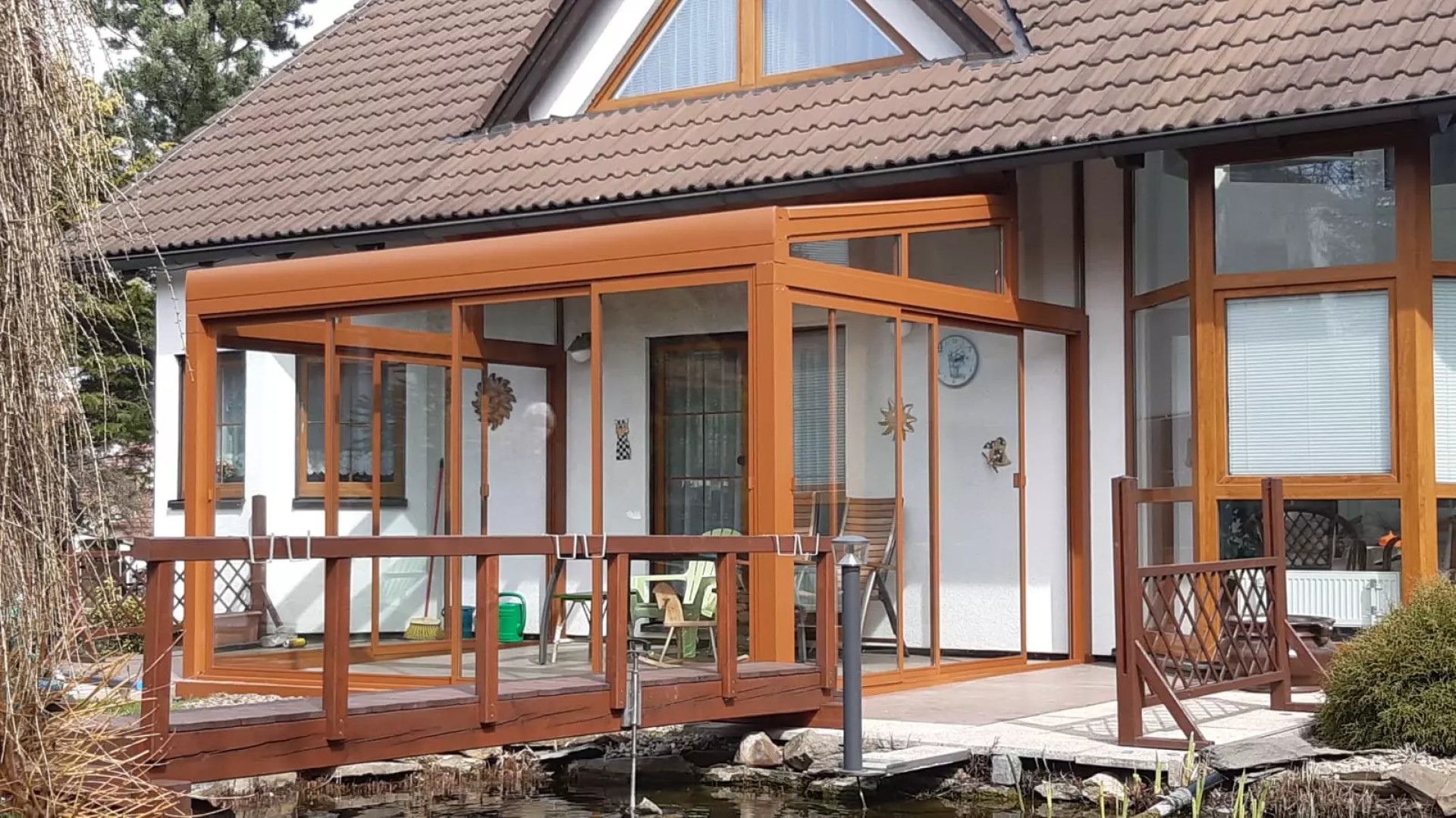 Die verglaste Aluminium-Pergola ROBUST in Braun neben einem Einfamilienhaus mit einer Holzbrücke
