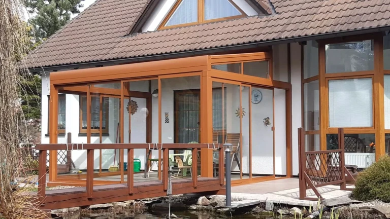 Die verglaste Aluminium-Pergola ROBUST in Braun neben einem Einfamilienhaus mit einer Holzbrücke