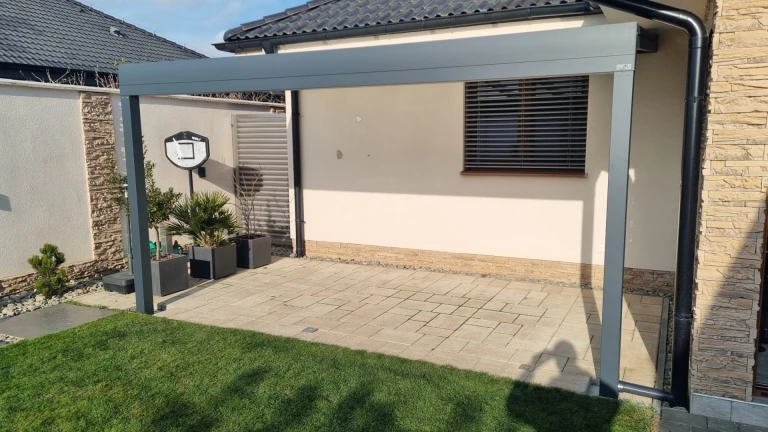Eine moderne graue Aluminium-Pergola über der Terrasse eines Einfamilienhauses mit Pflaster und Rasenfläche