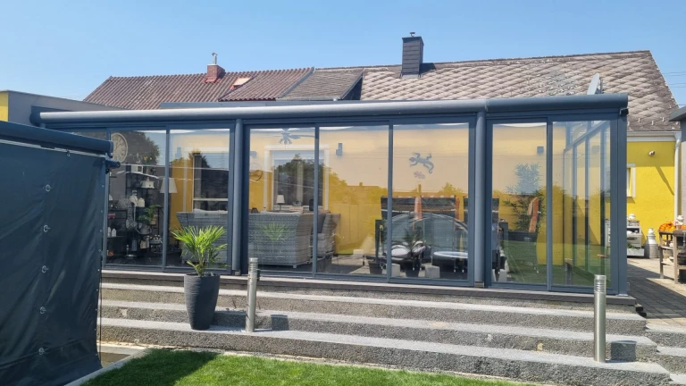 Ein moderner, verglaster Wintergarten aus Aluminium in Anthrazit, der an ein gelbes Einfamilienhaus mit Treppenhaus angebaut ist.