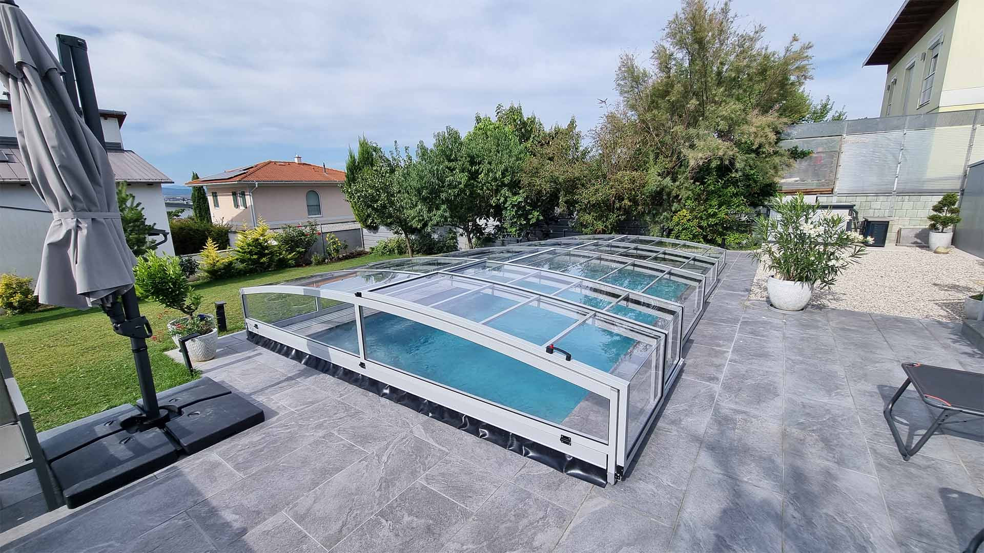 Moderne, niedrige Aluminium-Schwimmbadüberdachung vom Typ PREMIER auf der gepflasterten Terrasse im Garten eines Einfamilienhauses