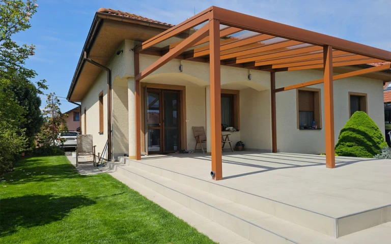 Eine moderne, braune Aluminium-Pergola mit Glasdach auf der gepflasterten Terrasse eines Einfamilienhauses