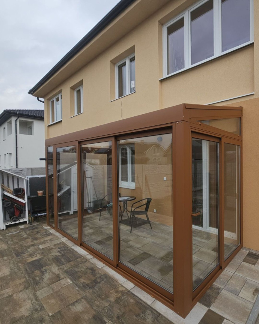 Ein moderner, verglaster Wintergarten mit brauner Konstruktion, der an ein Einfamilienhaus angebaut ist